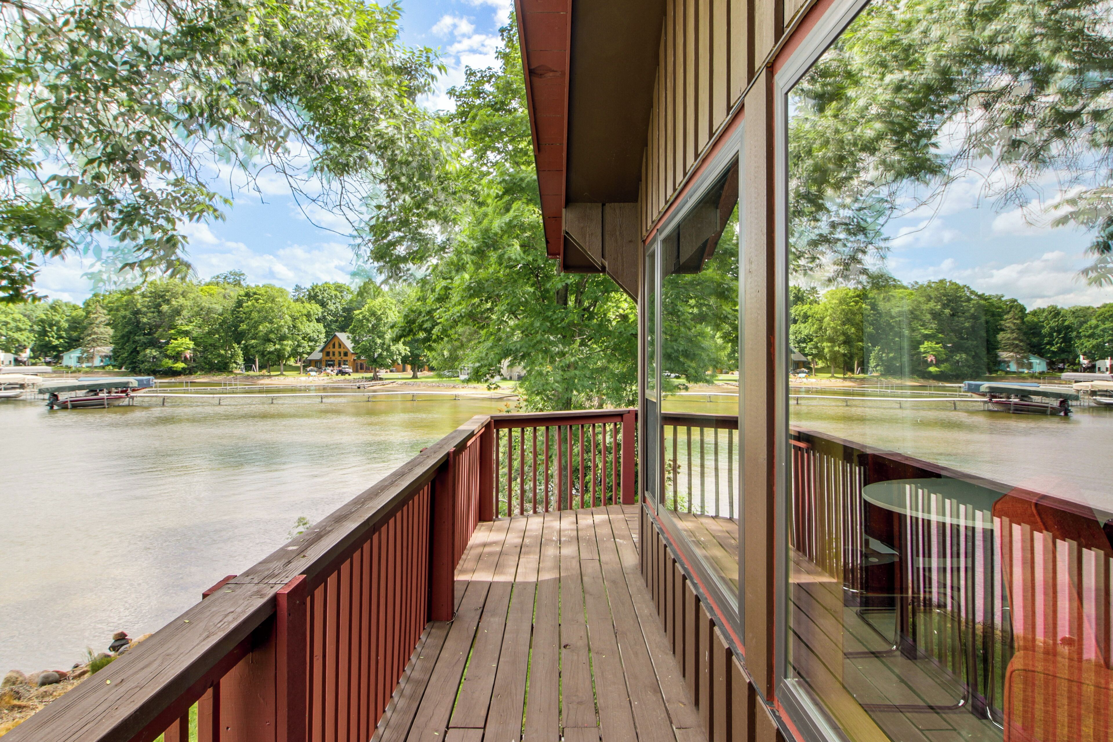 A wooden deck with a glass railing overlooking a calm lake, surrounded by trees.
