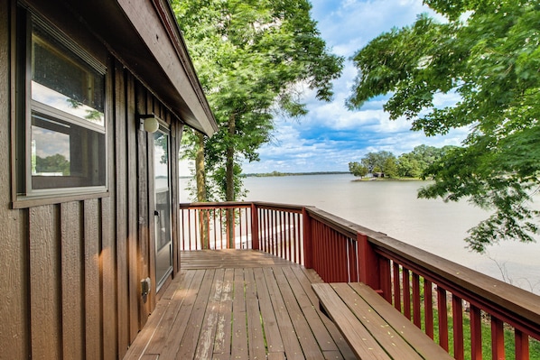 A wooden deck with a railing overlooking a lake, a building with large windows, and a clear sky.