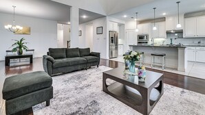A modern living room with a gray sofa, ottoman, and coffee table, a kitchen with white cabinets and stainless steel appliances, and a dining area with a bench and vase. - Near Lake & Family Fun! Spacious Oak Point Home (Little Elm)