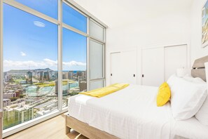 Une chambre avec une grande fenêtre, un lit avec une literie blanche et un oreiller jaune, et une vue sur la ville.
