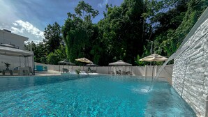 Ein klares Schwimmbecken mit Liegestühlen und Sonnenschirmen, umgeben von einer weißen Mauer und üppiger Bepflanzung.