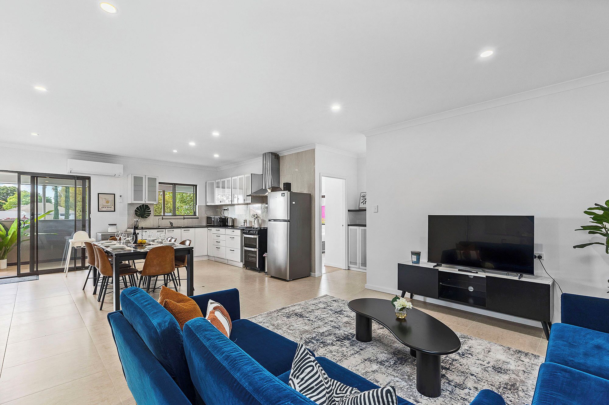 A modern living room with a sofa, coffee table, and TV. Adjacent is a kitchen with white cabinets and a dining area with a table set for four.