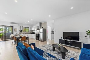 Een moderne woonkamer met een bank, salontafel en televisie. Grenzend aan de woonkamer bevindt zich een keuken met witte kasten en een eethoek met een tafel voor vier personen.