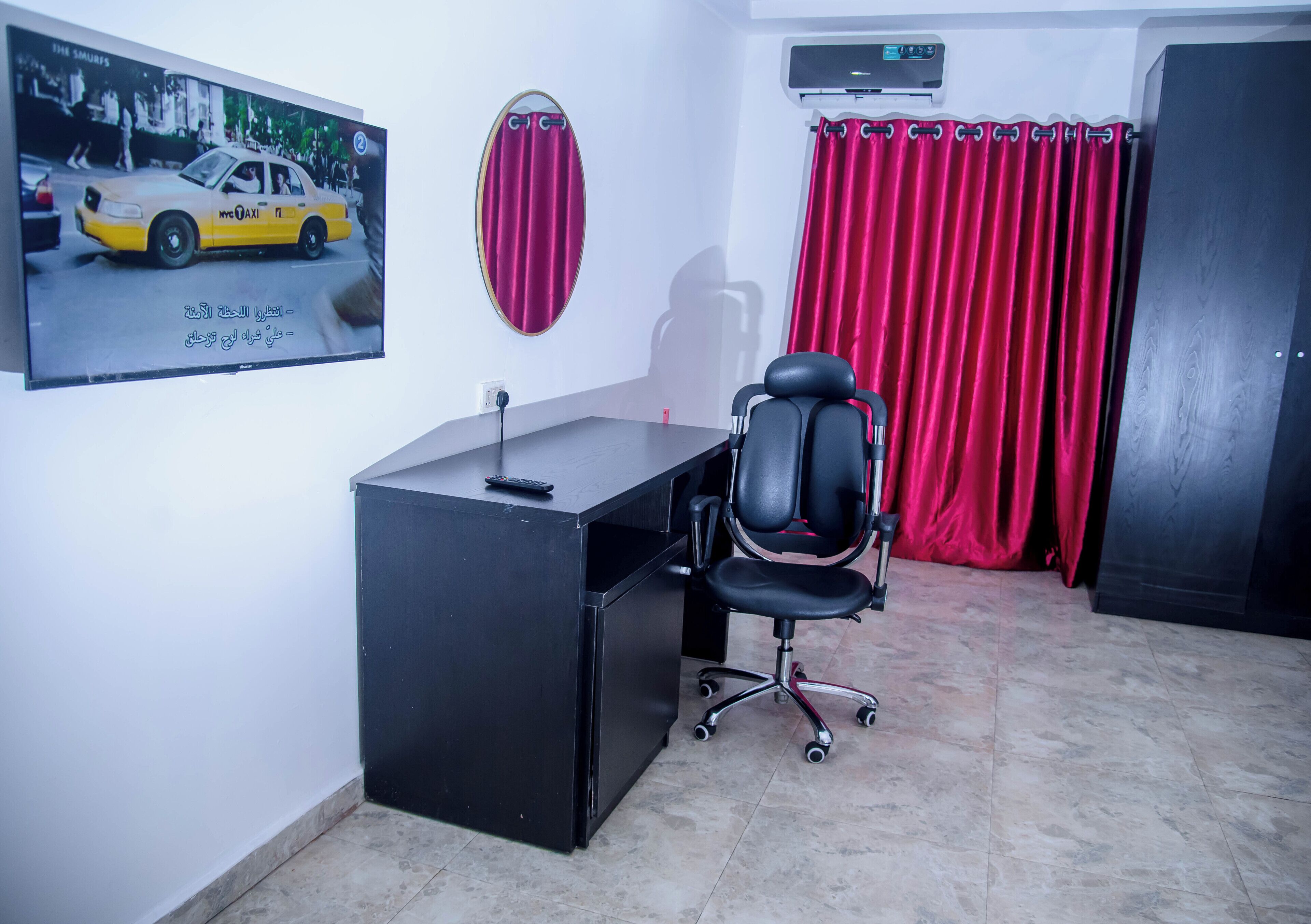 A room with a desk, a black office chair, a TV displaying a taxi, a mirror, and red curtains.