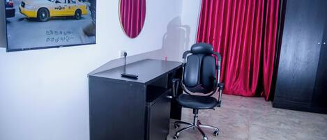 A room with a desk, a black office chair, a TV displaying a taxi, a mirror, and red curtains.