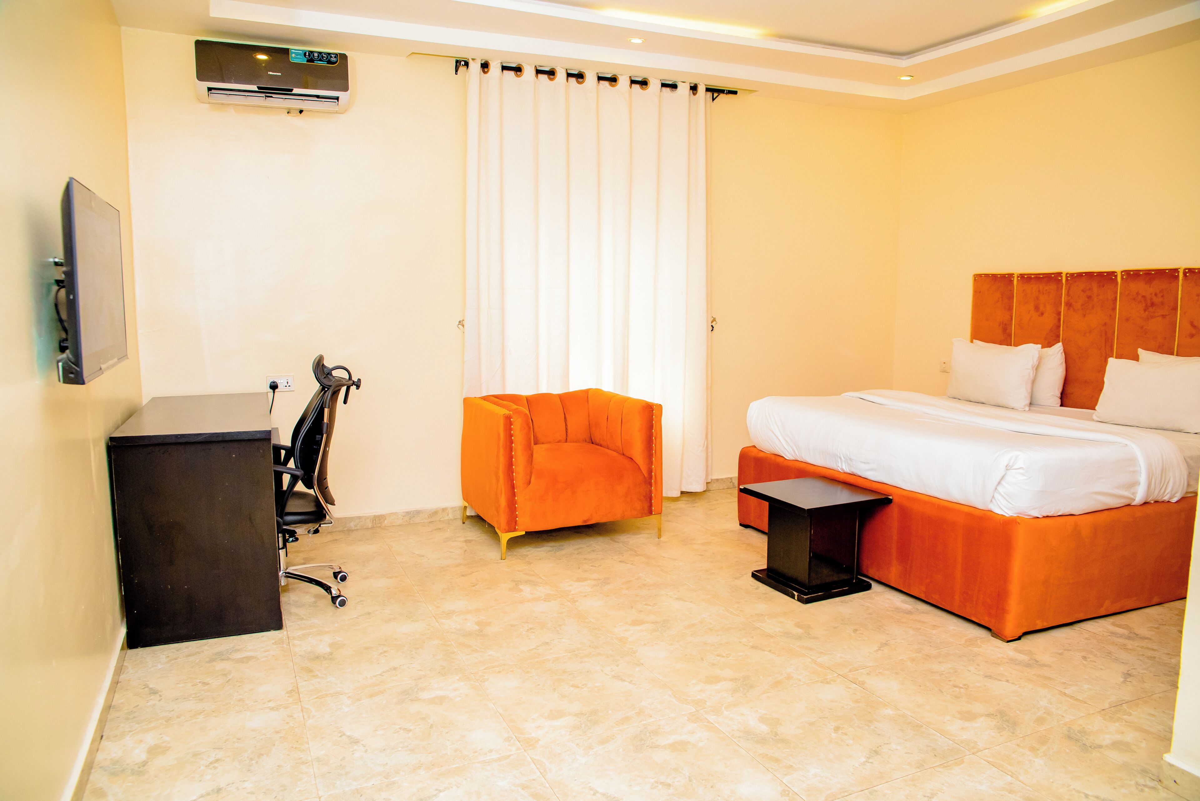 A hotel room with a bed, an orange chair, a desk, and an air conditioning unit.
