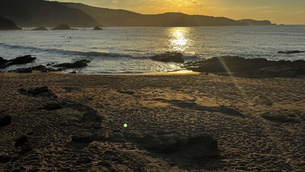 Ein Strand mit untergehender Sonne über dem Meer, felsiger Küste und Hügeln in der Ferne.