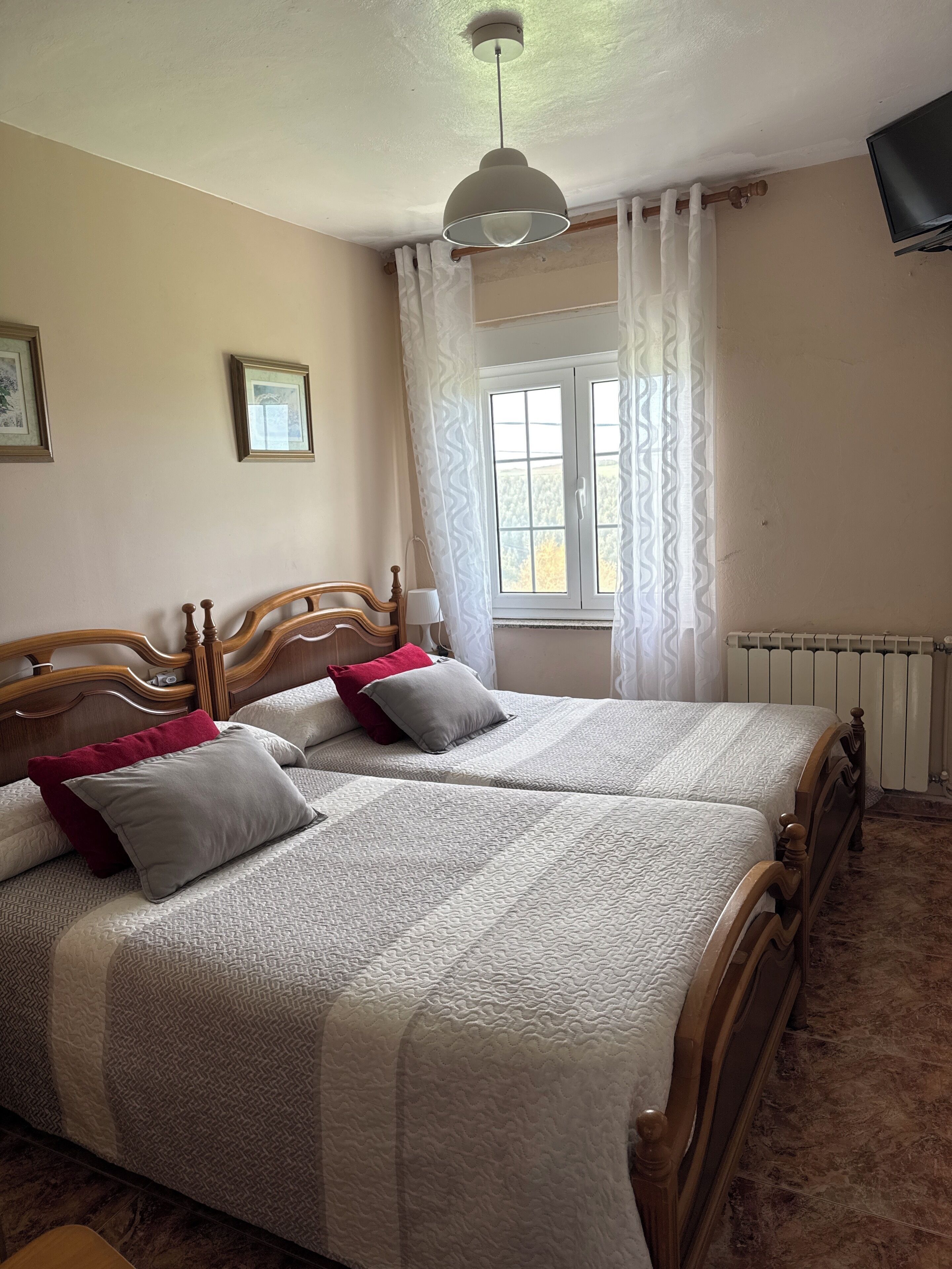 Ein Doppelbett mit Holz-Kopfteil, zwei Nachttische, ein Fenster mit Vorhängen, eine Deckenleuchte und ein an der Wand befestigtes Fernsehgerät.