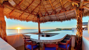 Área de bar com telhado de palha, com vista para um corpo d'água e um pôr do sol.