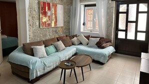 A living room with a sectional sofa, a coffee table, a painting on the wall, and a window with curtains. - Casa Joanna Old Town Rethymno (Rethymno)