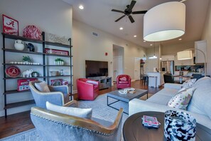 Uma sala de estar ampla com sofá, poltronas, mesa de centro e televisão. Cozinha visível ao fundo.