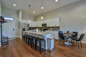 A modern kitchen with white cabinets, a central island with stools, a dining area with chairs, and pendant lighting.