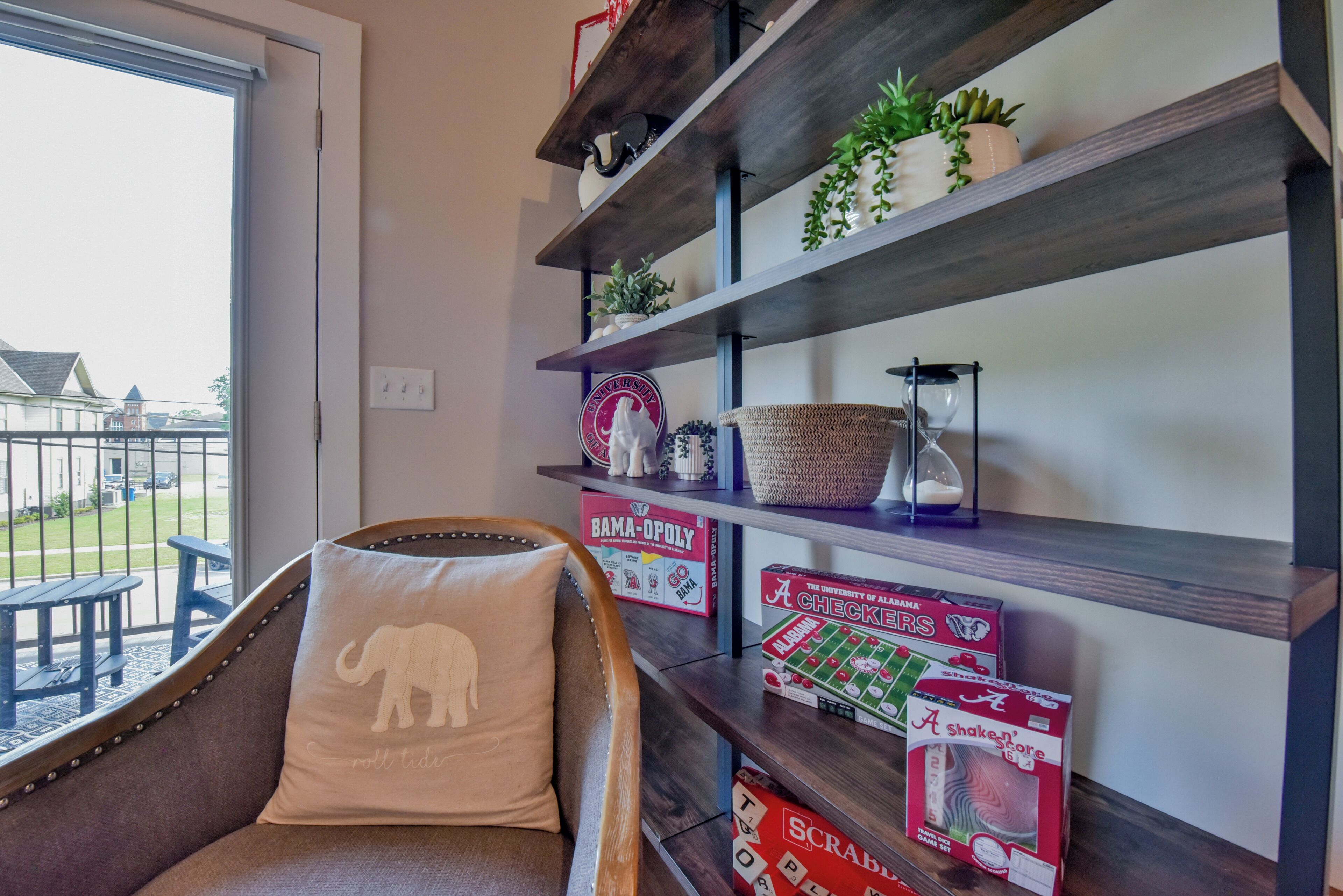 Een kamer met uitzicht op een balkon, een stoel met een decoratief kussen, en planken met diverse spullen, waaronder bordspellen en planten.