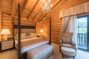 A four-poster bed with a tufted headboard, a nightstand with a lamp, a chair, and an ottoman in a log cabin room with a large window.