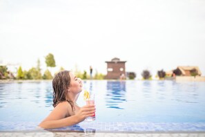 A person in a swimming pool with a drink, buildings in the background, and a clear sky.
