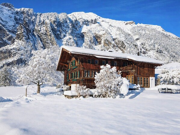 A wooden chalet with a sloped roof, surrounded by snow-covered trees and mountains.