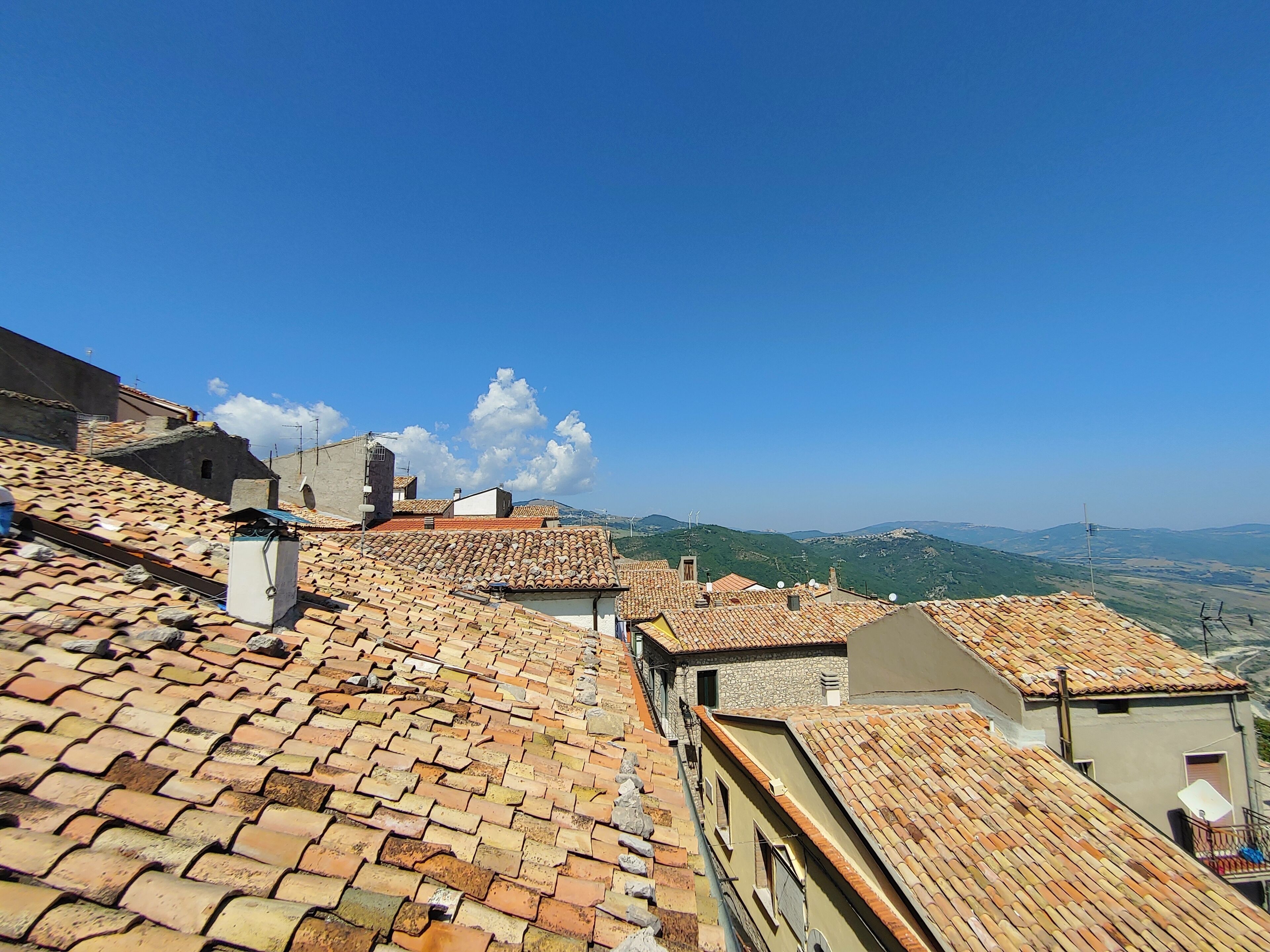 Una vista di tetti in tegole di terracotta, un edificio in pietra e un cielo azzurro con colline lontane.