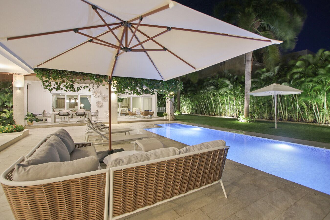 Un espace aménagé au bord de la piscine, avec un canapé en osier, un parasol et une vue sur une maison dotée d’une terrasse.