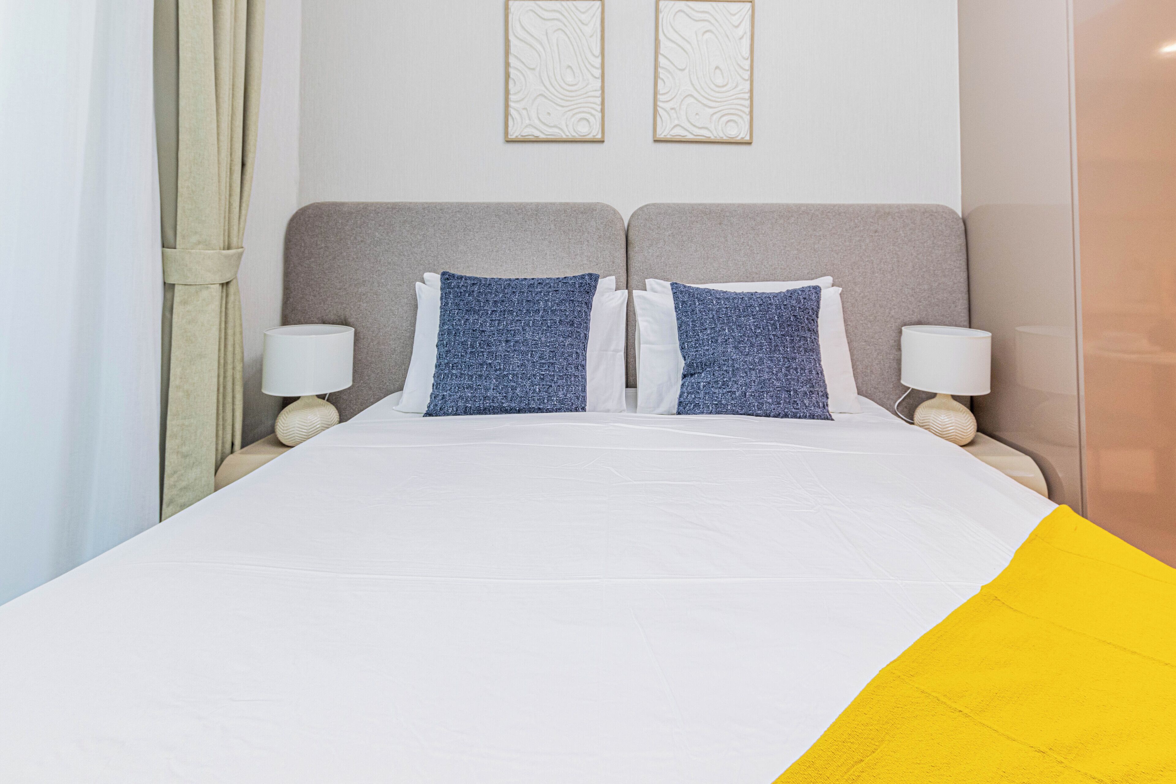 A double bed with white and blue bedding, two wall-mounted lamps, and two framed wall art pieces.