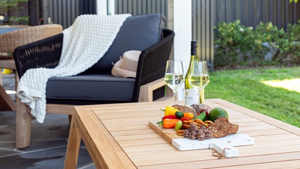 Een houten tafel met diverse etenswaren en twee glazen witte wijn, gelegen op een terras met een rieten stoel en een witte deken.