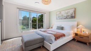 Een slaapkamer met een groot raam, een bed met witte beddengoed, een nachtkastje met een lamp en een kunstwerk aan de muur.