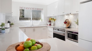 A modern kitchen with white cabinetry, a large window, a sink, an oven, and a bowl of fruit on the counter. - Sunshine Retreat – relaxing coastal retreat (Culburra Beach)