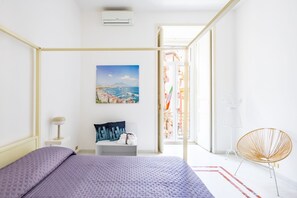 Ein Schlafzimmer mit Bett, Nachttisch, Sessel, Gemälde und Blick ins Freie.