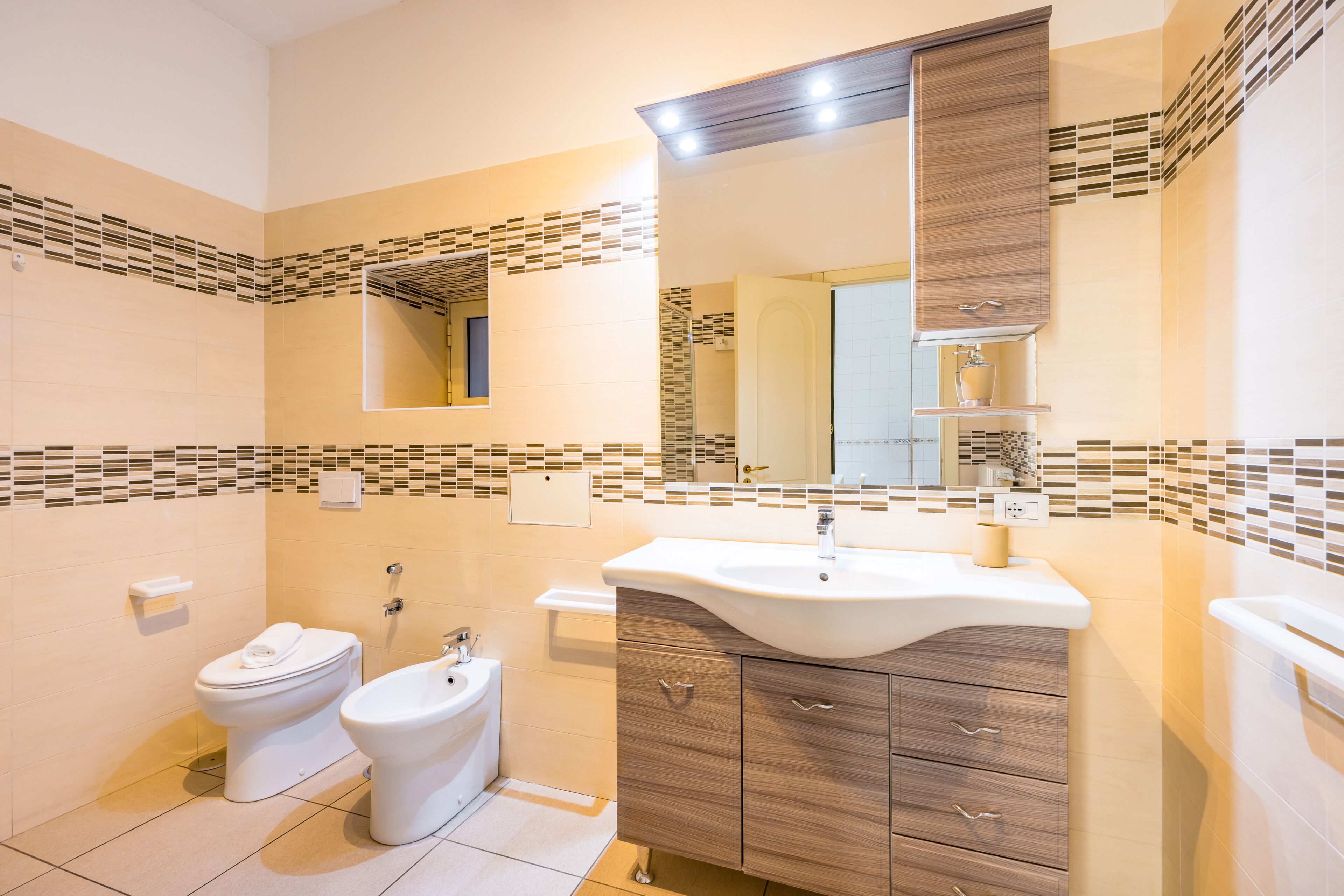 Een moderne badkamer met toilet, bidet, wastafel met spiegel en betegelde wanden.