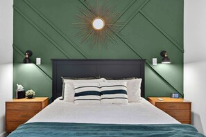 Una cama bien tendida con un cabecero oscuro, dos mesitas de noche con lámparas y un espejo con forma de sol montado en la pared.