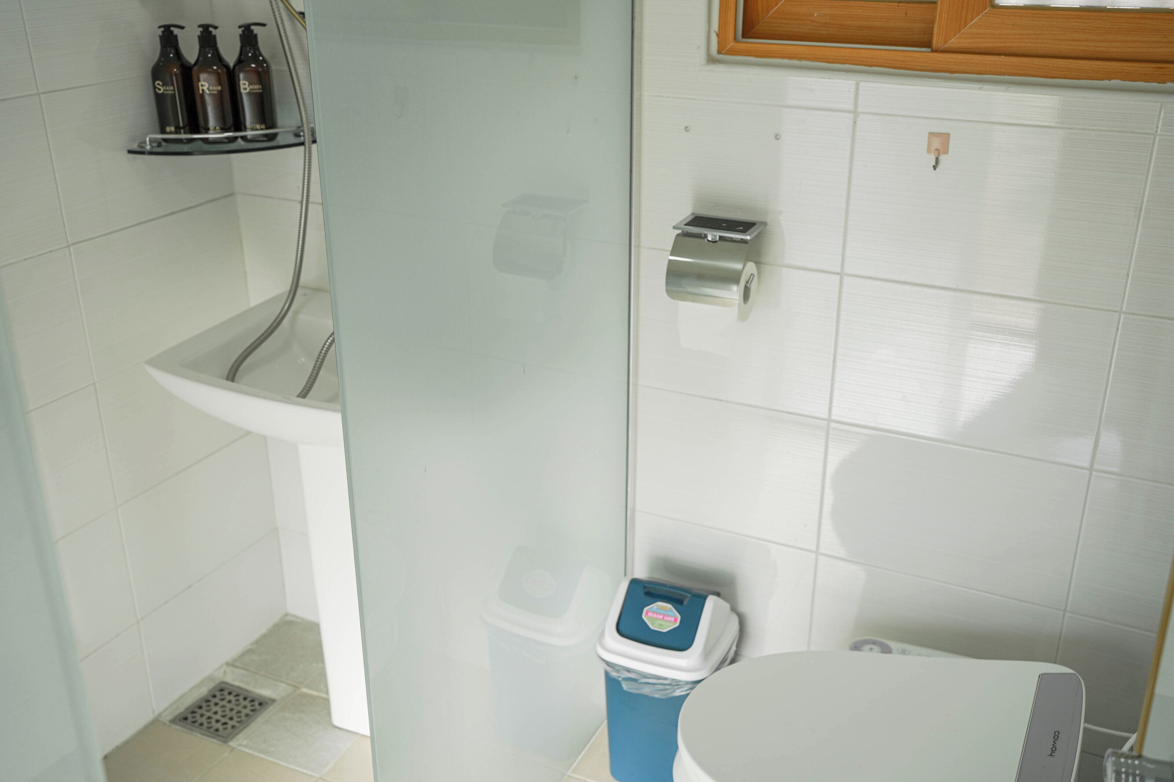A bathroom with a toilet, a trash bin, a soap dispenser shelf, and a shower area.