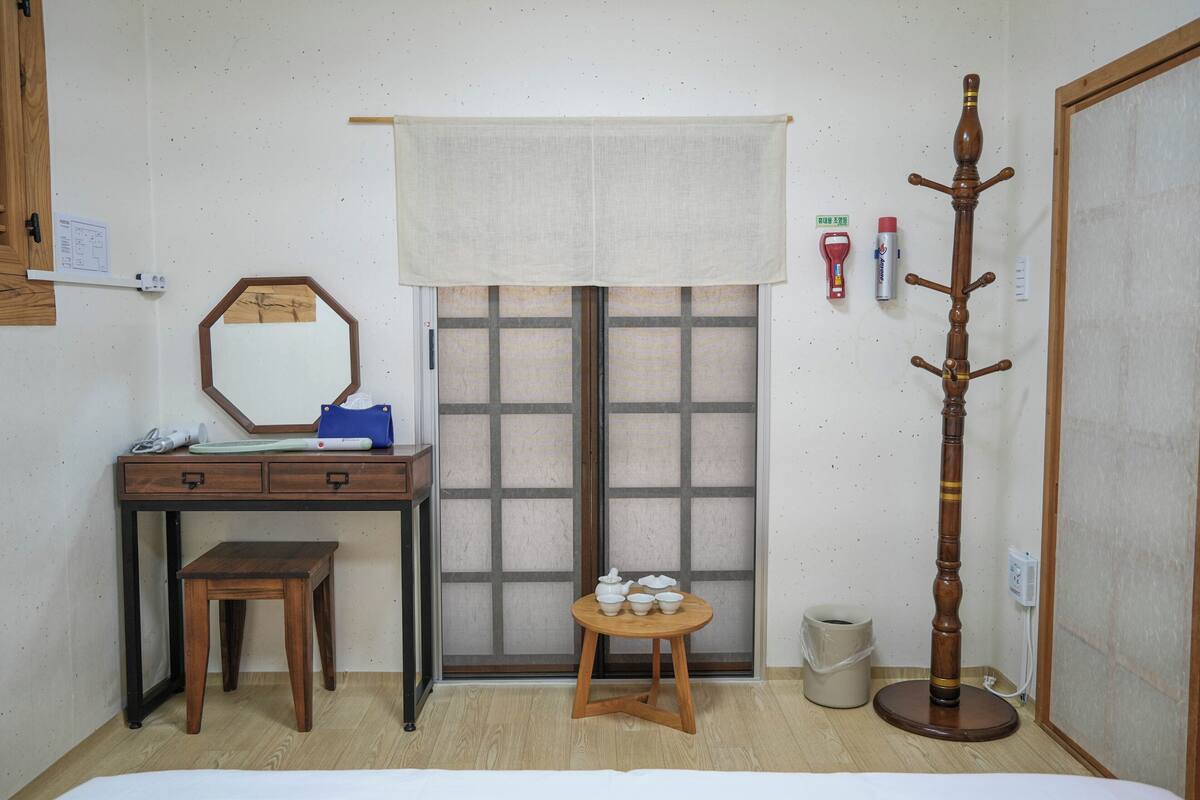 Een kamer met een houten bureau, een klein krukje, een spiegel, een raam met gordijn, een kapstok en een prullenbak.