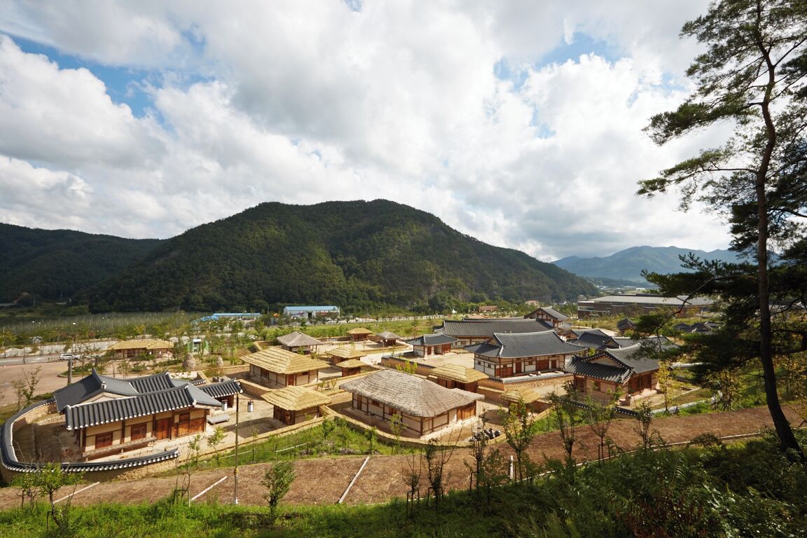 A traditional Korean village with wooden houses, a curved building, and mountains in the background.