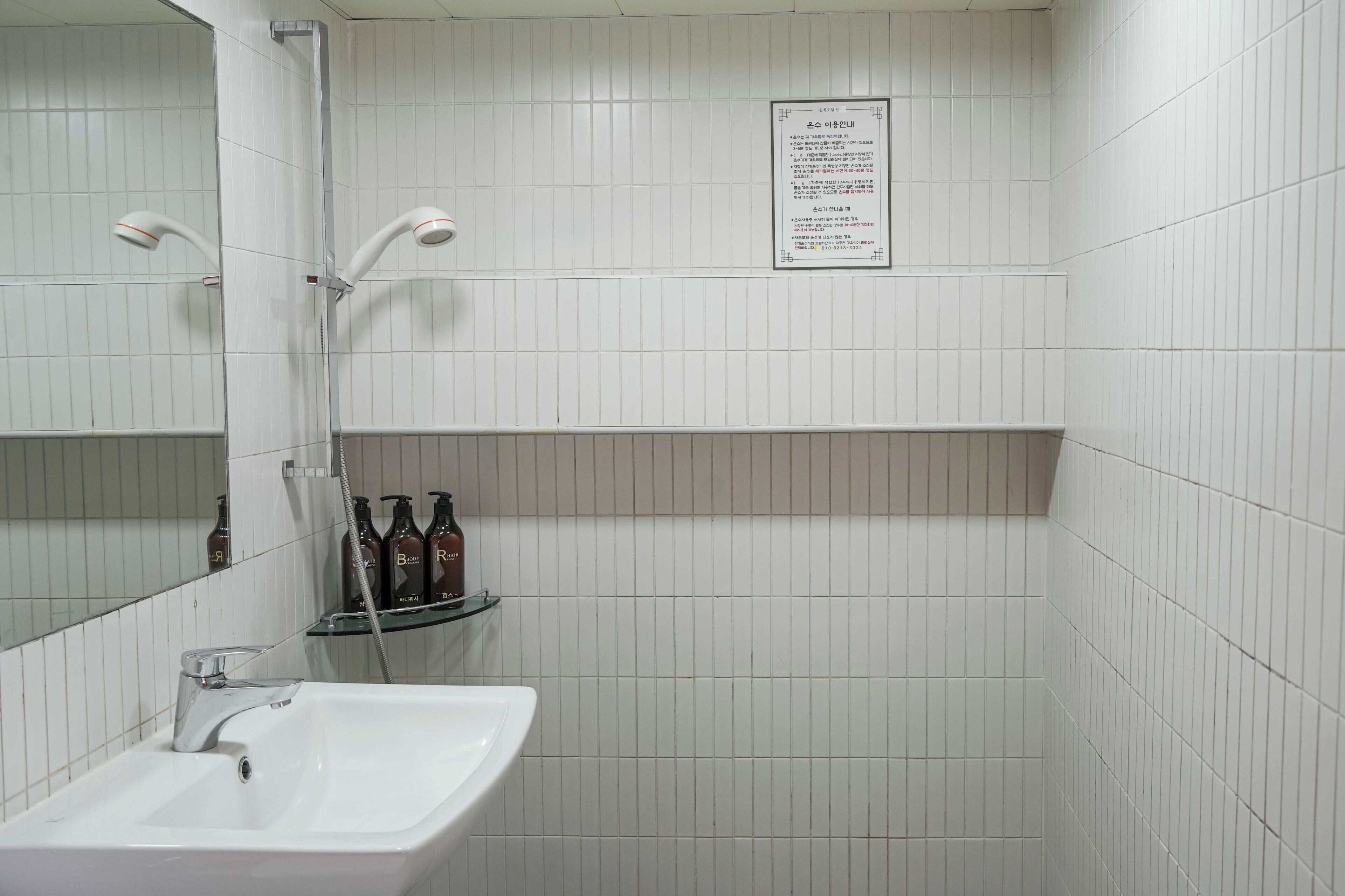 A bathroom with a sink, mirror, shower, and a shelf with bottles.