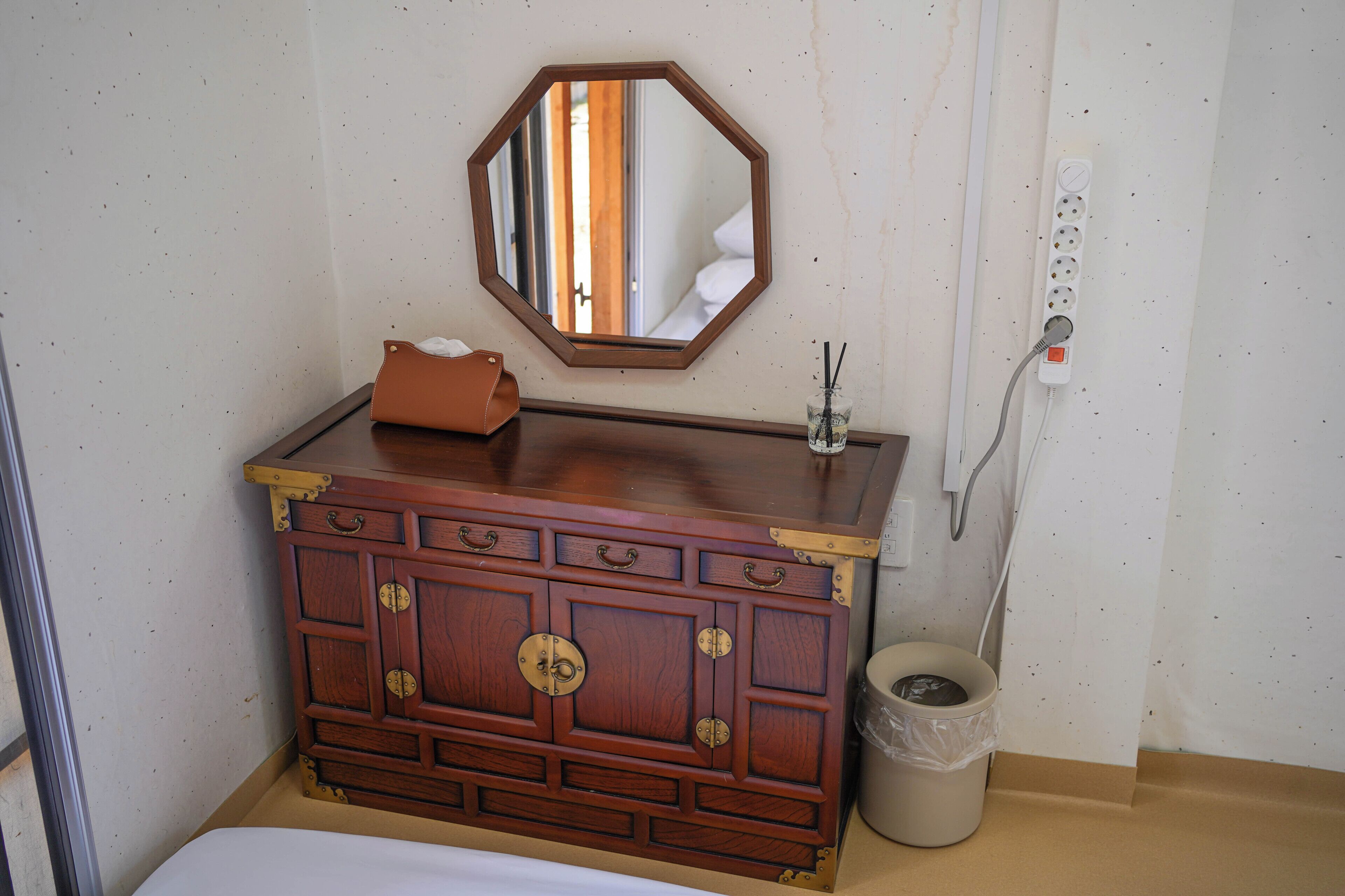A wooden dresser with drawers and a mirror, a trash bin, and a power outlet on the wall.