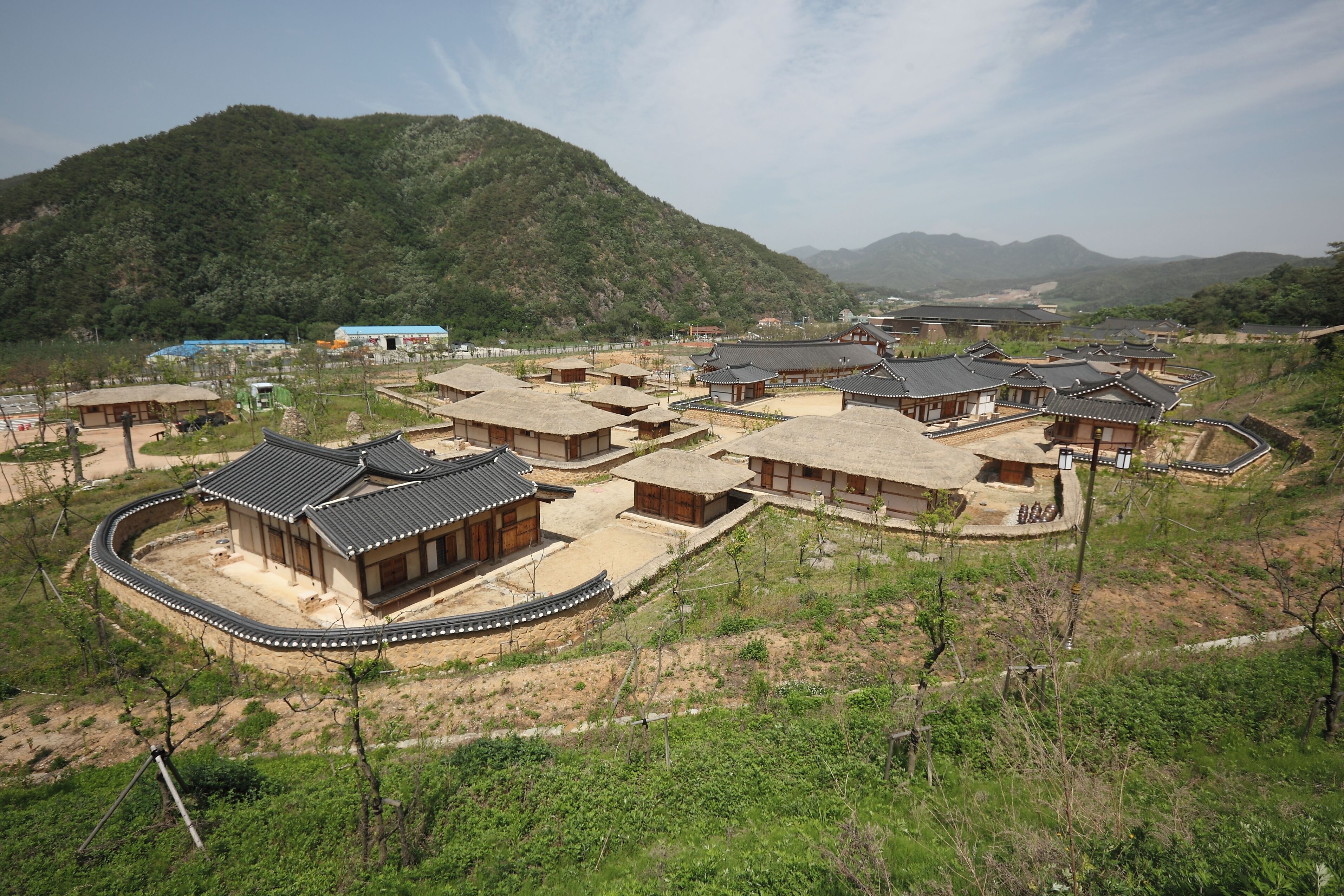 A traditional village with multiple buildings, some with curved roofs and others with flat roofs, surrounded by greenery and hills.