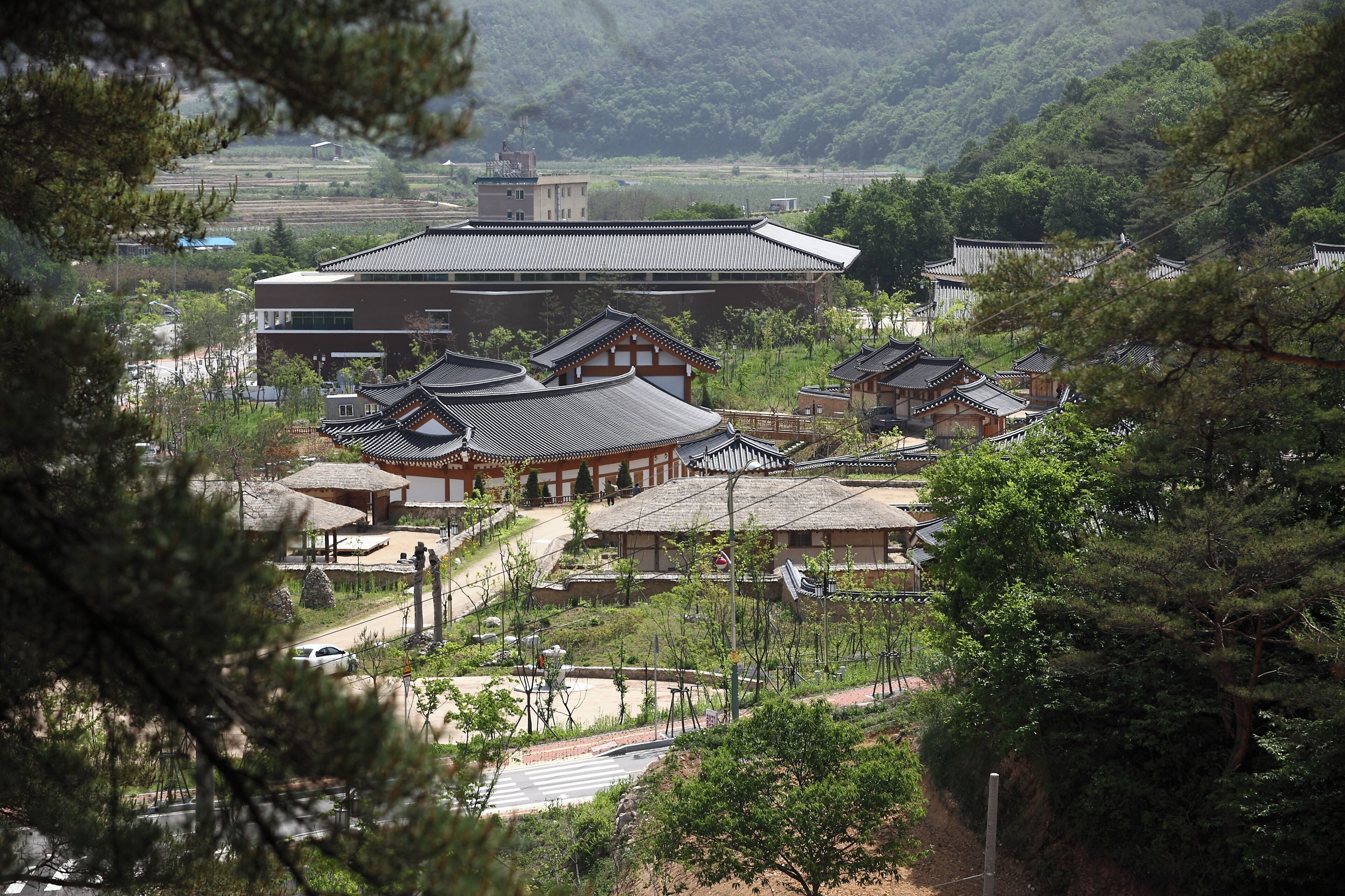 A traditional Korean hanok-style building complex with multiple structures, surrounded by lush greenery and a hilly landscape.
