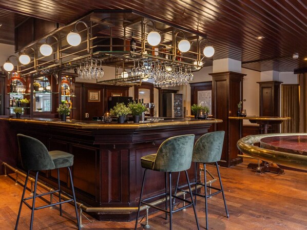 A bar area with a wooden bar, three bar stools, a chandelier, and a fish tank.
