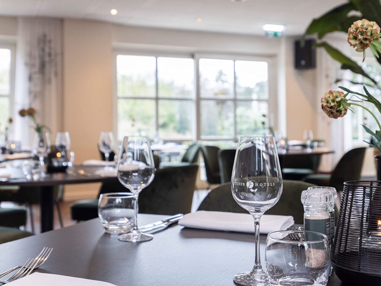 A dining table set with wine glasses, a salt and pepper shaker, and a menu stand in a restaurant with large windows and a view of greenery.