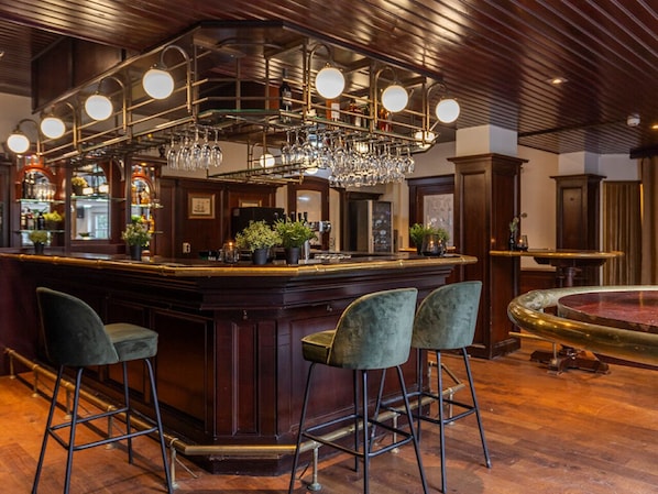 A bar area with a wooden bar, three bar stools, a chandelier, and a fish tank.