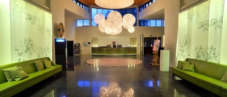 En rummelig lobby med en central reception, grønne sofaer, en stor lysekrone og et højt loft med træbjælker.