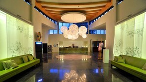 A spacious lobby with a central reception desk, green sofas, a large chandelier, and a high ceiling with wooden beams.
