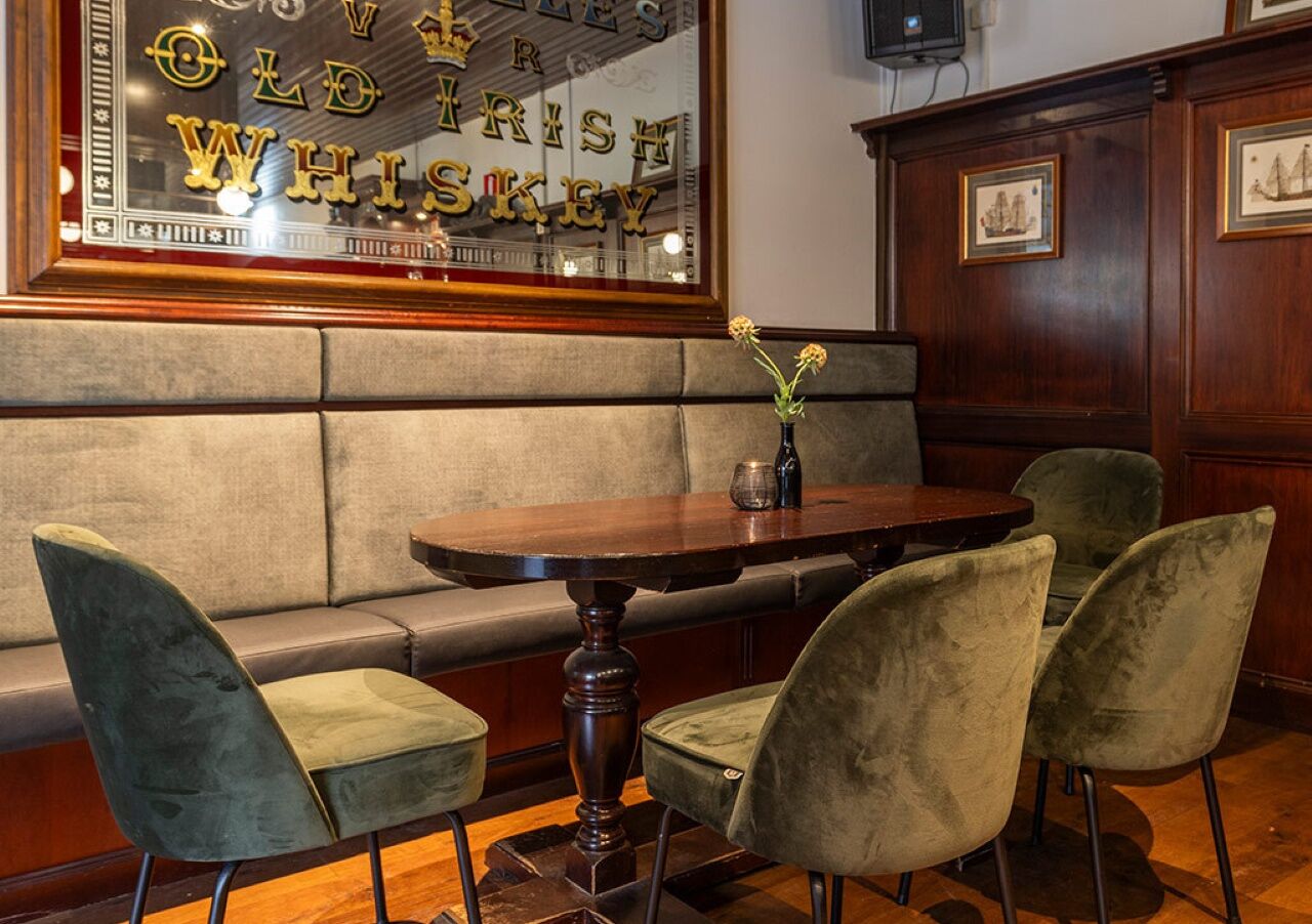 Un coin salon accueillant avec une table ronde en bois, des chaises rembourrées et un miroir décoratif portant l’inscription « Old Irish Whiskey ».