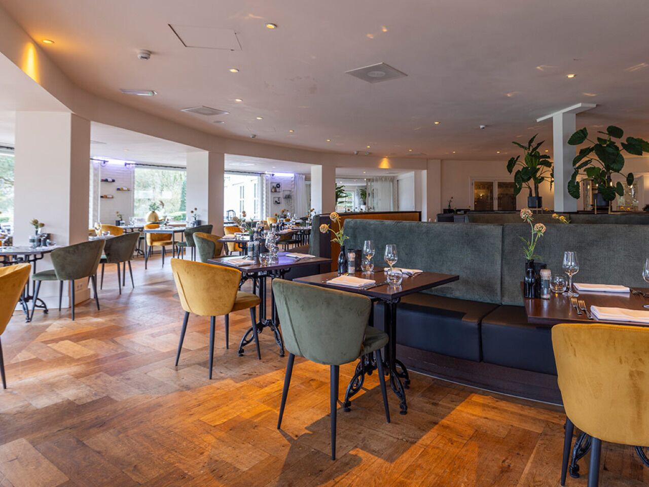 Ein modernes Restaurant mit Holzböden, mehreren Sitzbereichen inklusive Tische und Sitzecken, sowie großen Fenstern für viel Tageslicht.