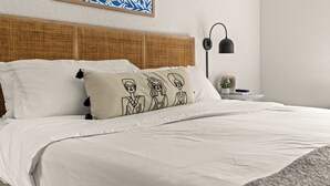 Een netjes opgemaakt bed met witte lakens, een rieten hoofdbord en een kussen met een tekening van drie personen.