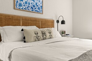 Ein ordentlich bezogenes Bett mit weißer Bettwäsche, einem Rattan-Kopfstück und einem Kopfkissen mit einer stilisierten Zeichnung von drei Personen.
