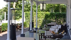 Une terrasse extérieure avec des tables et des chaises, un vase décoratif rempli de bouteilles, et une haie verdoyante en arrière-plan.