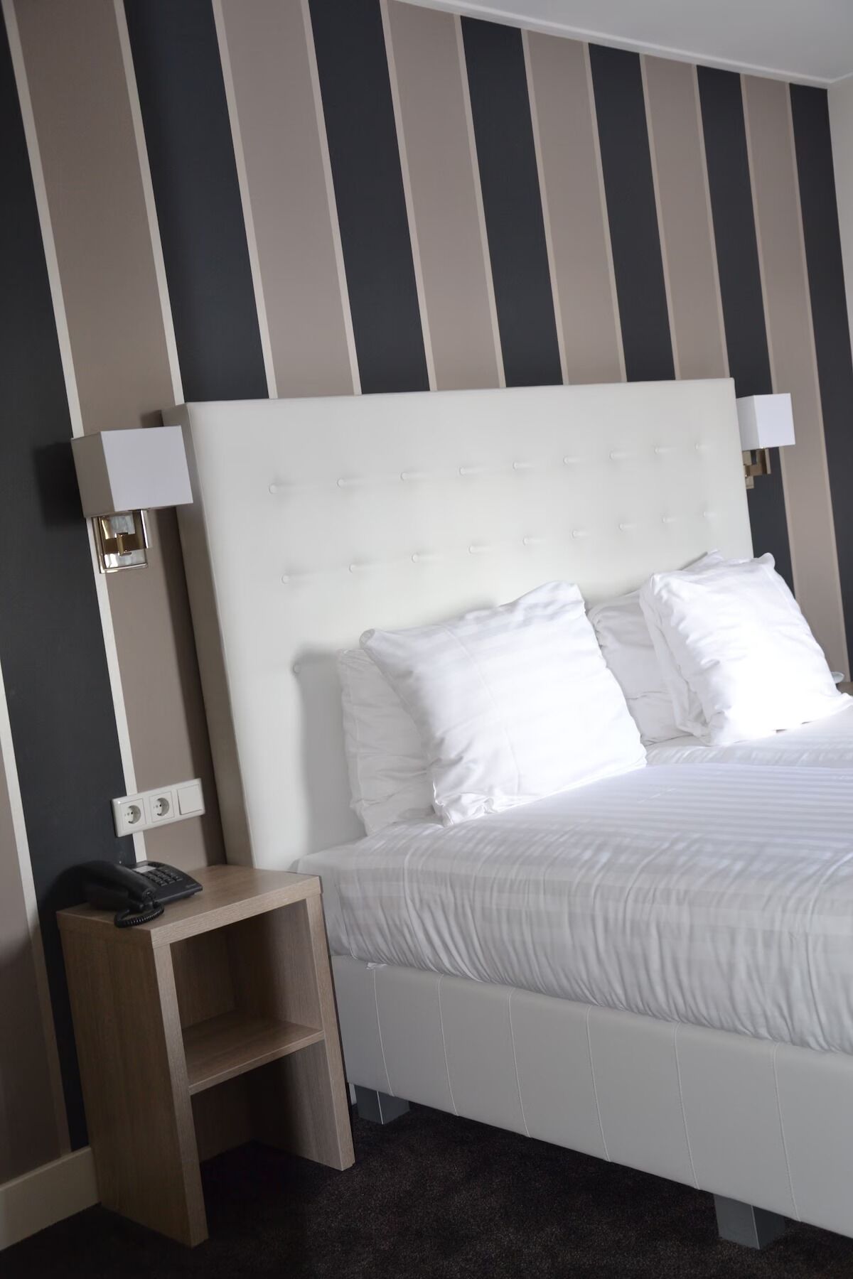 A bed with white bedding, a bedside table with a phone, and a wall with vertical stripes.