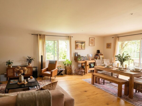Un salon comprenant un canapé, une table basse en bois, une table à manger pour quatre personnes et une chaise. On y trouve également des fenêtres avec des rideaux, une bibliothèque et un ventilateur.