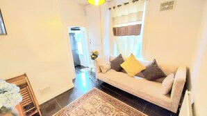 A living room with a sofa, a small table with flowers, a rug, and a kitchen in the background.