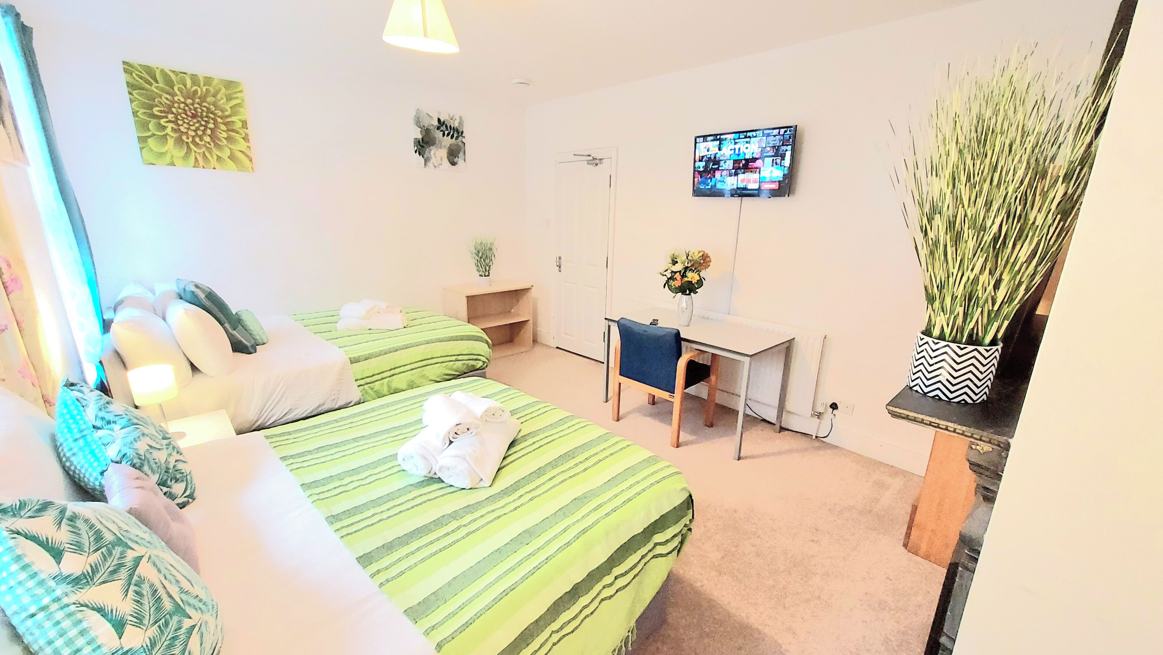 A hotel room with two beds, a desk, a chair, a TV, and a potted plant.